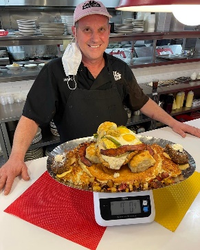 worker standing behind a counter; before him is a tray of what looks like breakfast foods, on a scale; it weighs 7 lbs