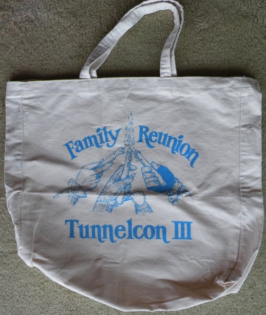 tote bag, white canvas printed in blue; image is 3 hands holding candles coming together in one flame; a furry baby hand holds the baby finger of Vincent's hand; text above the image: Family Reunion; text below: Tunnelcon III